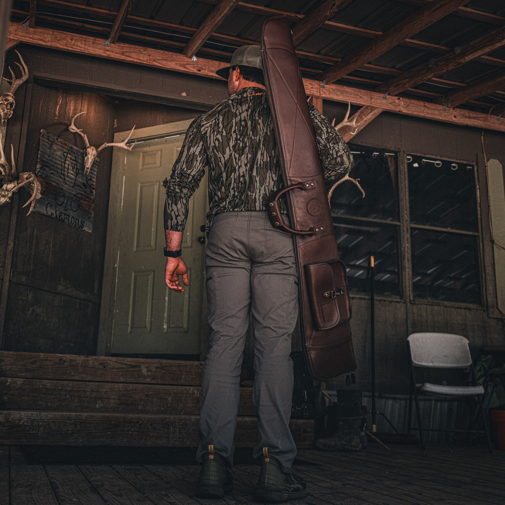 Man in camouflage holding a large leather gun case in a rustic cabin setting.